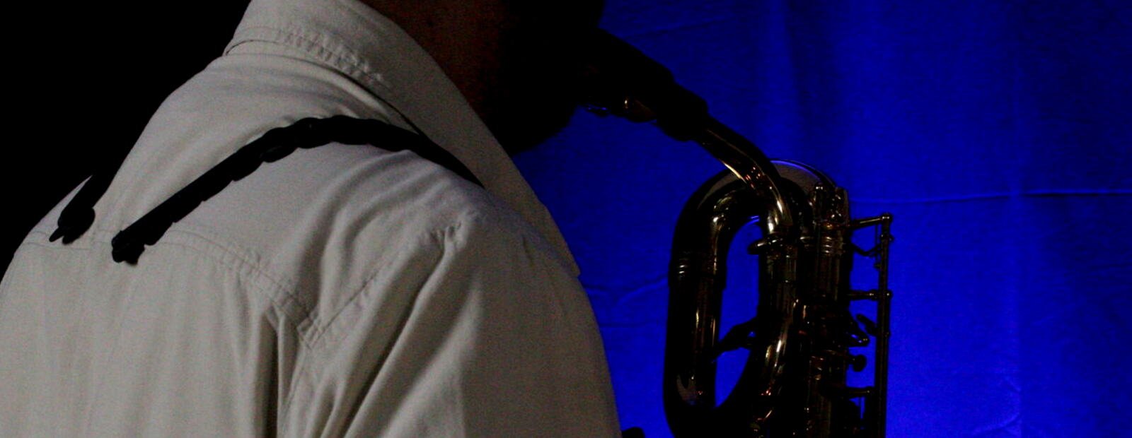 Ash Belgeri from behind, baritone saxophone in hand, blue backlight